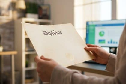 Anonymous hands gently hold a French diploma with a subtle seal. Blurred modern office background with a digital screen suggests new opportunities.