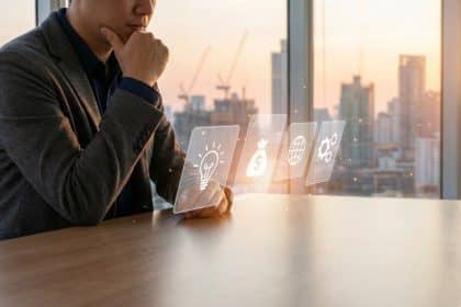 Man contemplating entrepreneurial ideas (lightbulb, money, globe, gear) at a modern desk with a city skyline at dawn.
