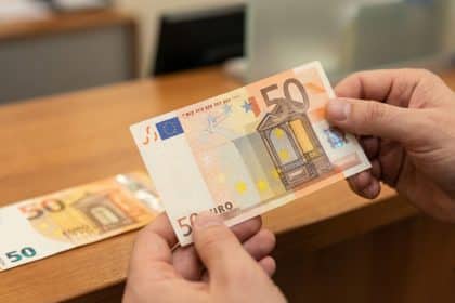 Close-up of hands holding a crisp Europa series 50 euro banknote, with an older 50 euro note blurred on a wooden counter.