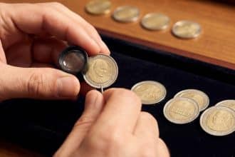 Close-up of hands examining a 2002 Vatican 2 euro coin with a jeweler's loupe, surrounded by other rare coins.
