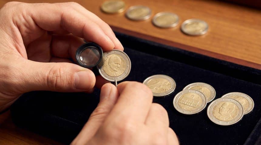 Close-up of hands examining a 2002 Vatican 2 euro coin with a jeweler's loupe, surrounded by other rare coins.