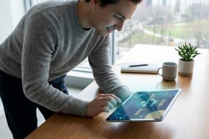 Un homme souriant se penche pour interagir avec une tablette posée sur une table en bois, affichant une grille de puzzle numérique avec des éléments futuristes. Vue sur une ville.