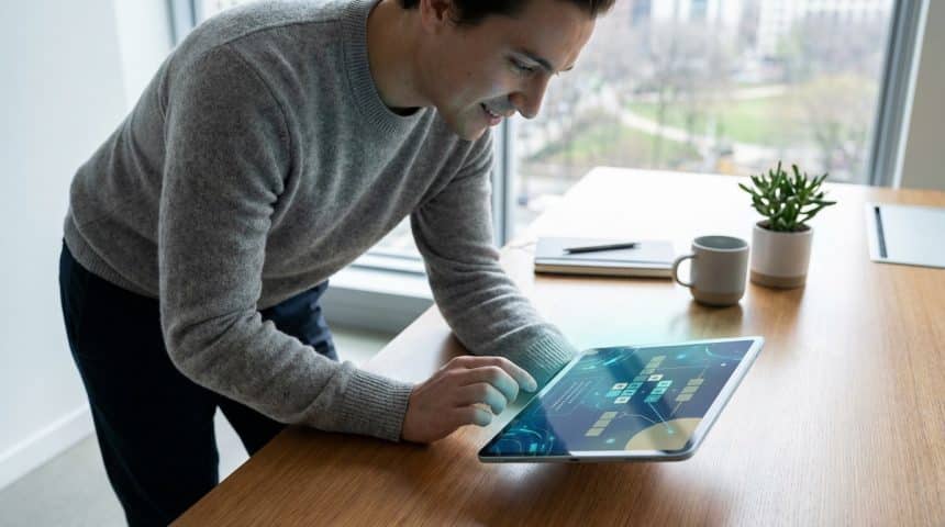 Un homme souriant se penche pour interagir avec une tablette posée sur une table en bois, affichant une grille de puzzle numérique avec des éléments futuristes. Vue sur une ville.