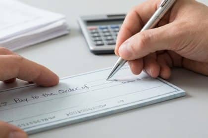 Vue rapprochée d'une personne tenant un stylo et écrivant sur un chèque vierge. Une calculatrice et des papiers sont en arrière-plan.