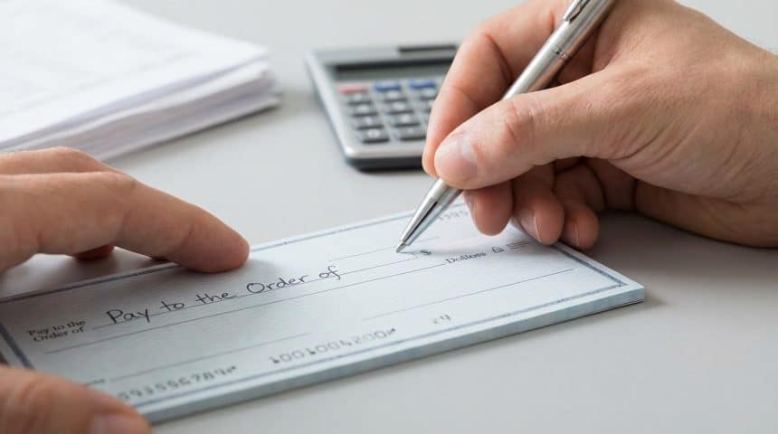 Vue rapprochée d'une personne tenant un stylo et écrivant sur un chèque vierge. Une calculatrice et des papiers sont en arrière-plan.