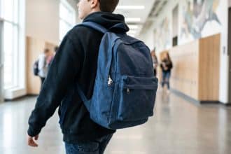 Jeune collégien de dos avec un sac à dos bleu. Il marche dans un couloir d'école avec des casiers et d'autres élèves.