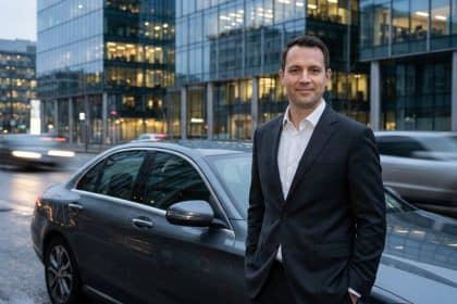 Homme professionnel souriant en costume, debout à côté d'une voiture grise de luxe en ville, avec des bâtiments modernes en arrière-plan et des voitures floues en mouvement.